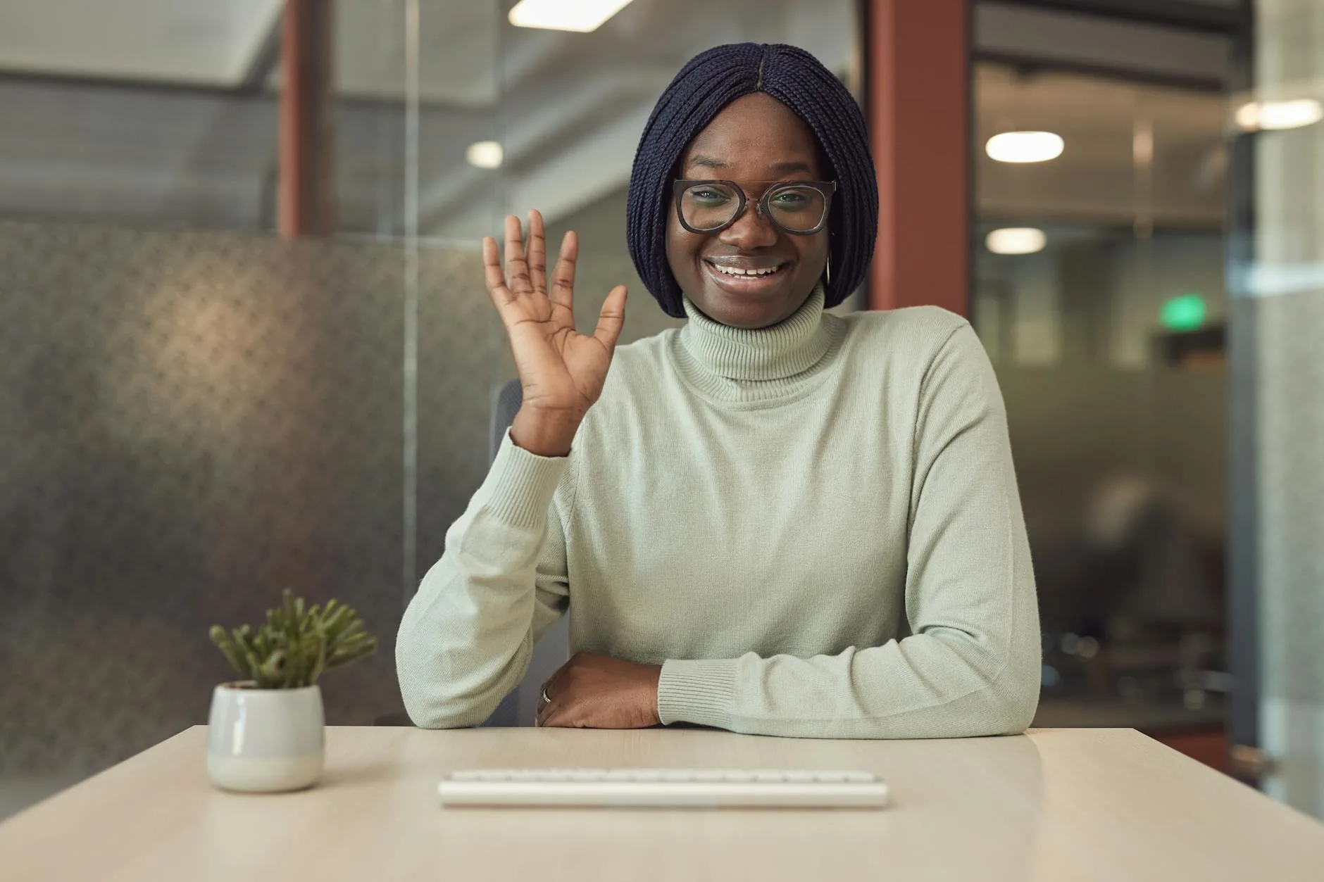 Professional African American woman smiling and waving in a modern office setting, showcasing friendliness and confidence.