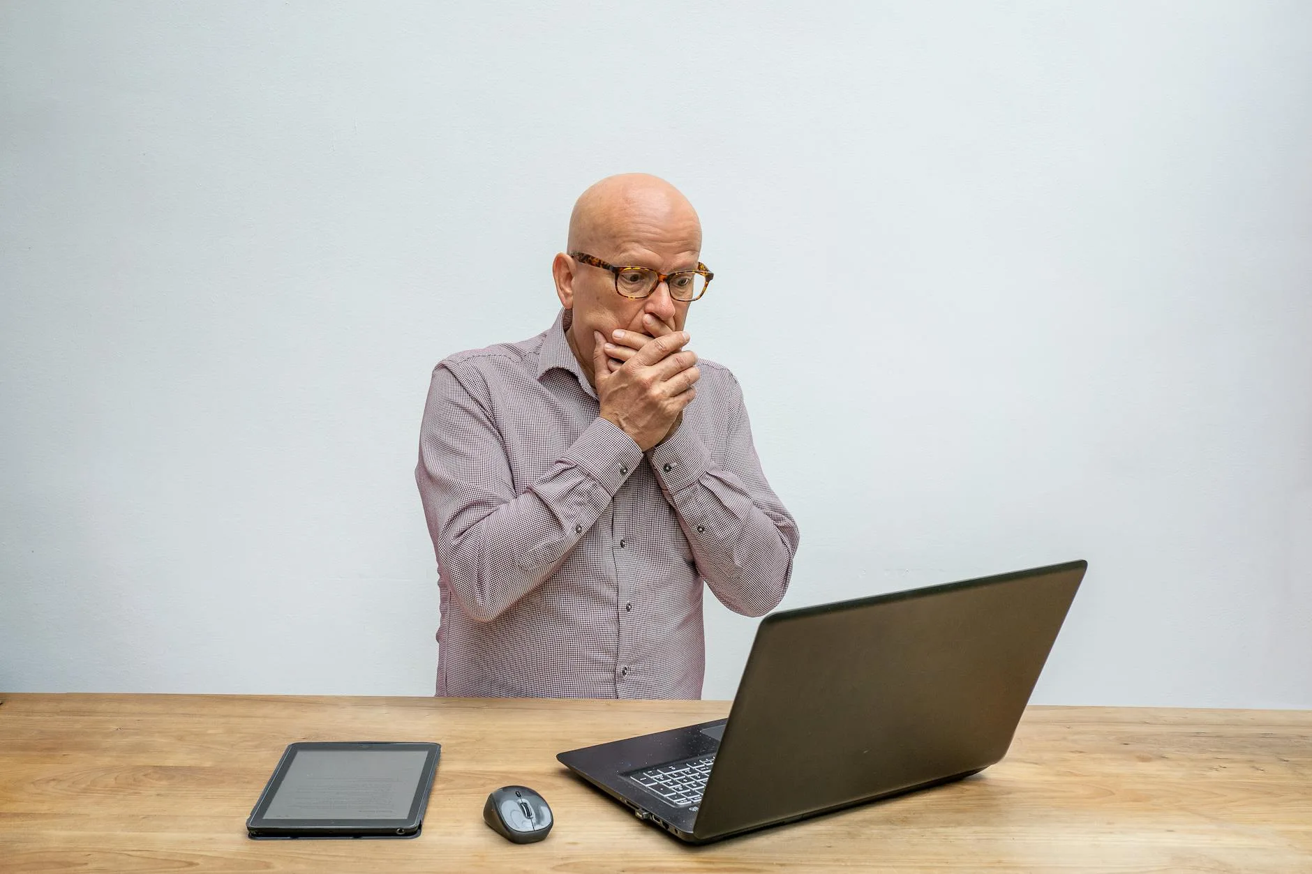 Elderly man reacts in surprise to laptop screen at wooden desk with tablet and mouse.