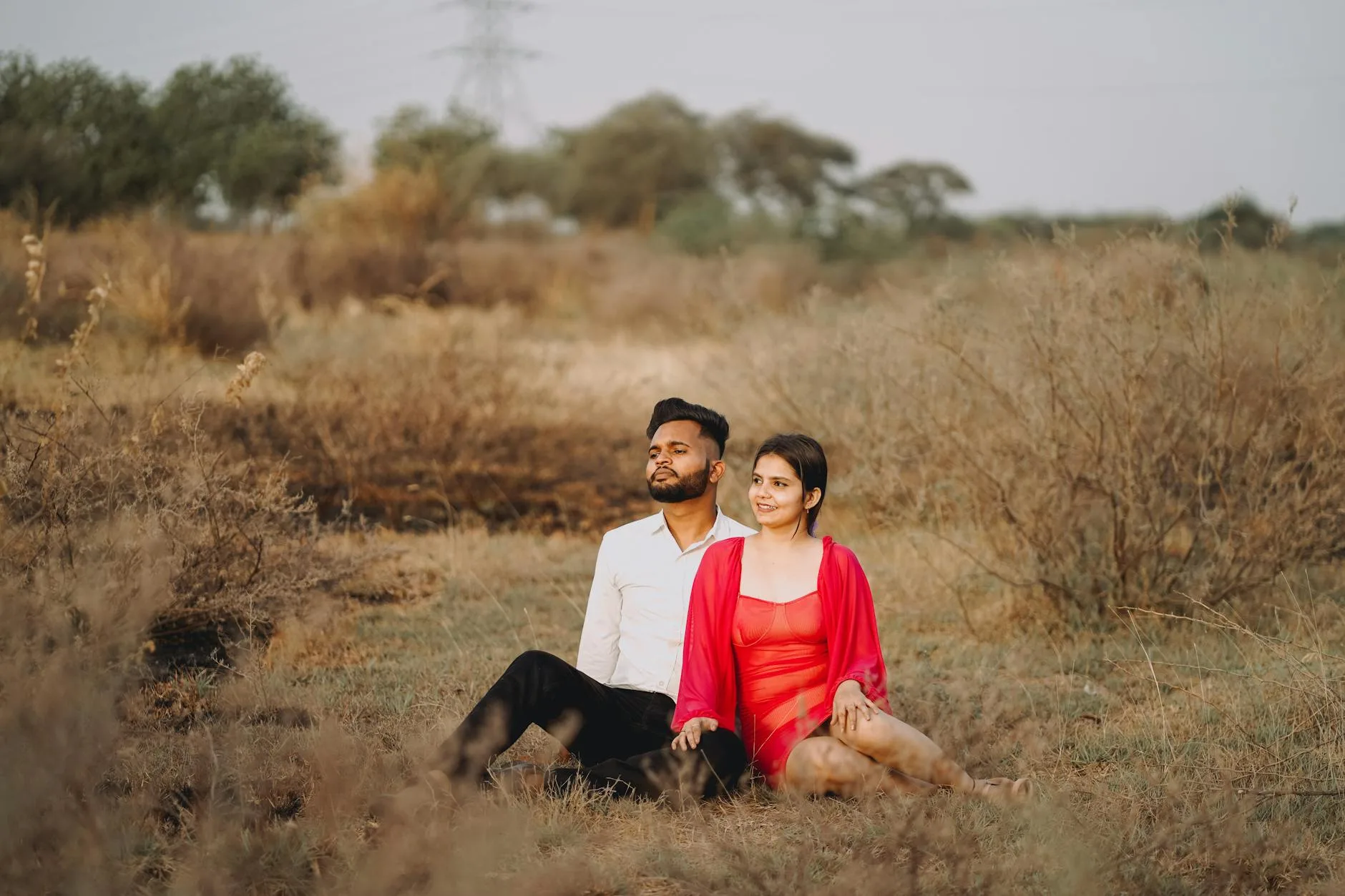 Happy couple sitting on dry grass, enjoying leisure time in serene countryside setting.
