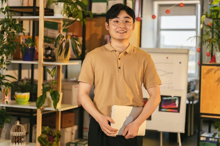 A smiling adult man with a laptop in a stylish office surrounded by plants.